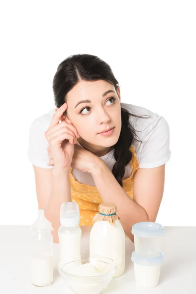 Jolie femme dans tablier avec doigt sur le temple à table avec récipients en plastique, bol, bouteille de lait et biberons — Photo de stock