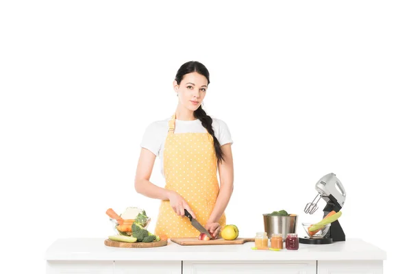 Jeune femme dans tablier coupe pomme à table avec mélangeur, légumes, bocaux avec purée et casserole — Photo de stock