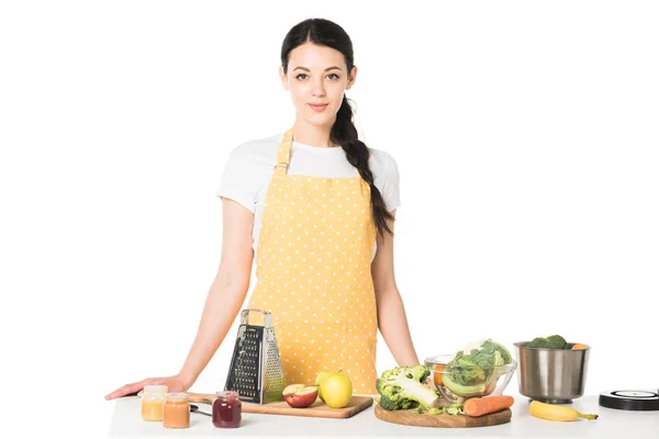 Mujer en delantal de pie en la mesa con frutas, verduras, tablas de cortar, rallador, frascos con puré y cacerola - foto de stock