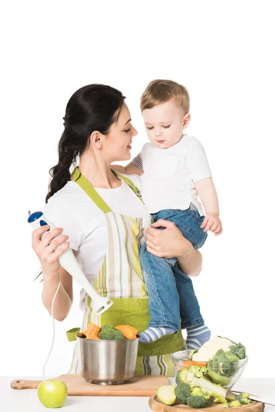 Smiling mother holding son and hand blender at table with apple and vegetables isolated on white background — Stock Photo