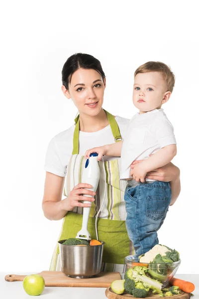 Mãe segurando liquidificador de mão e filho em pé na mesa com ingredientes isolados no fundo branco — Fotografia de Stock