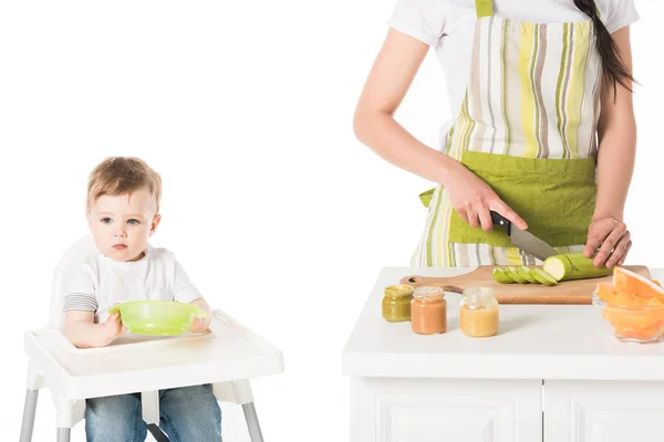 Cropped shot of mother in apron cutting zucchini and son sitting in highchair with plate — Stock Photo