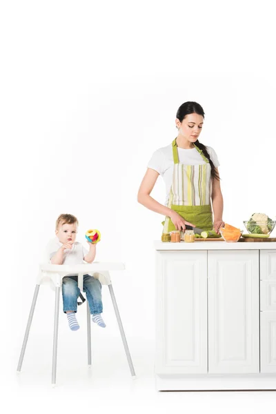 Baby Junge im Hochstuhl mit Spielzeug und Mutter schneiden Zucchini am Tisch isoliert auf weißem Hintergrund — Stockfoto