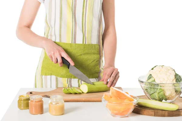 Cropped shot of woman cutting zucchini by knife at table with puree jars, cauliflower and pumpkin — Stock Photo