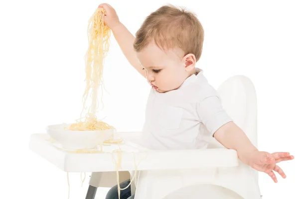 Baby boy holding spaghetti in hands and sitting in highchair isolated on white background — Stock Photo