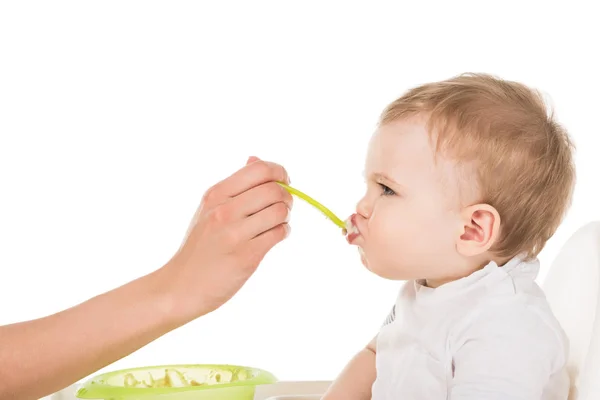 Imagen recortada de madre alimentando adorable pequeño hijo en trona aislado sobre fondo blanco - foto de stock