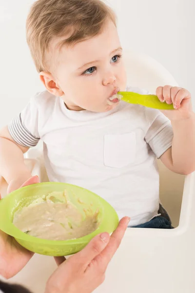 Recortado tiro de madre sosteniendo plato con comida y bebé niño con cuchara en silla alta - foto de stock