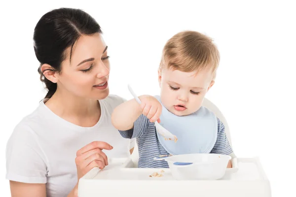 Mujer joven con hijo en babero comiendo y sentado silla alta aislado sobre fondo blanco - foto de stock