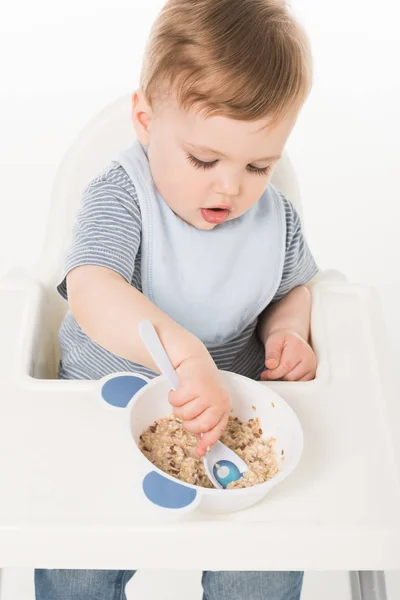 High angle view of baby boy in bib eating and sitting highchair isolated on white background — Stock Photo