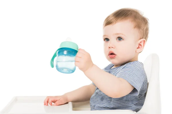 Adorável menino segurando copo de bebê com água e sentado em cadeira alta isolado no fundo branco — Fotografia de Stock