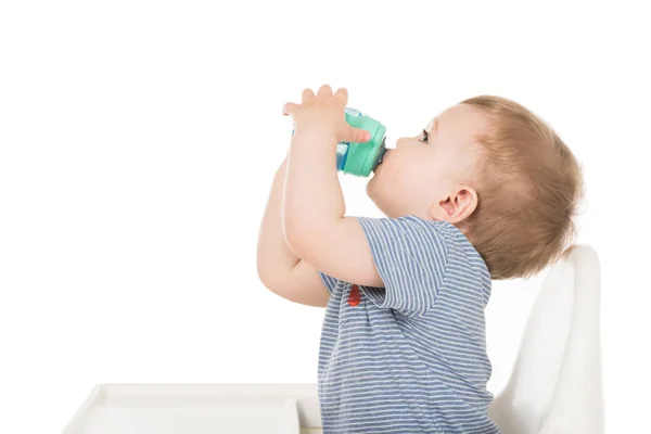 Vista lateral do menino bebendo água do copo do bebê e sentado em cadeira alta isolado no fundo branco — Fotografia de Stock