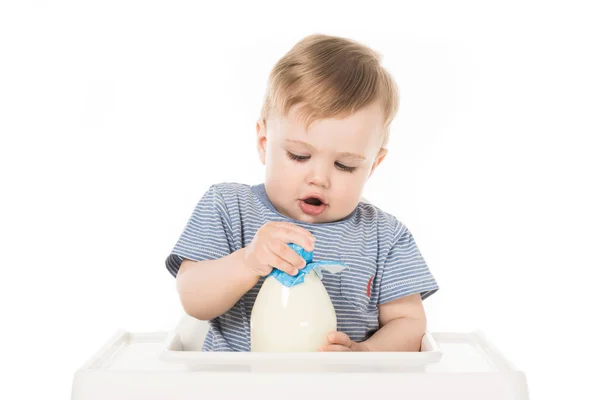 Маленький хлопчик намагається відкрити пляшку молока і сидить у високому кріслі ізольовано на білому тлі — стокове фото