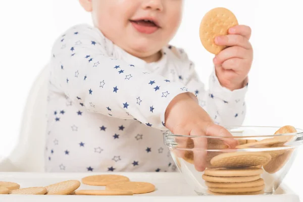 Abgeschnittene Aufnahme eines kleinen Jungen, der Kekse aus der Schüssel nimmt und isoliert im Hochstuhl auf weißem Hintergrund sitzt — Stockfoto