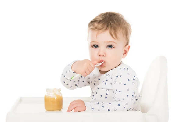 Adorable bebé niño comiendo puré de tarro y sentado en trona aislado sobre fondo blanco - foto de stock
