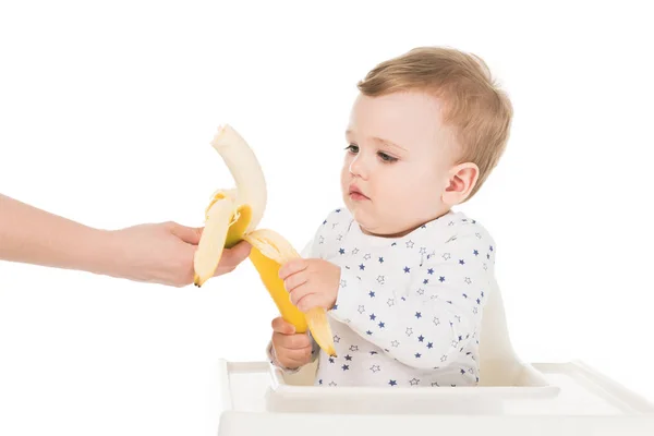 Recortado disparo de madre dando plátano a hijo en trona aislado sobre fondo blanco - foto de stock