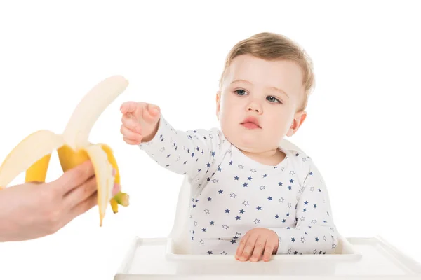 Imagen recortada de madre dando plátano a hijo en trona aislada sobre fondo blanco - foto de stock