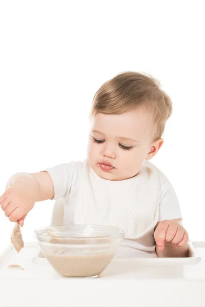 Bebé niño comiendo gachas por cuchara y sentado en trona aislado sobre fondo blanco - foto de stock