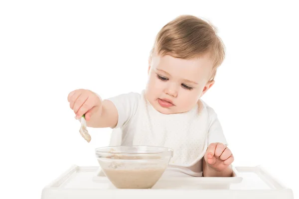 Niño en babero comiendo comida de bebé por cuchara y sentado en trona aislado sobre fondo blanco - foto de stock