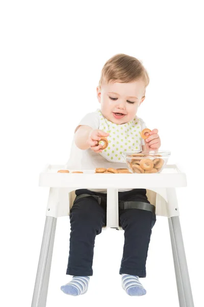 Lächelnder kleiner Junge mit Bagels und Schale sitzt isoliert im Hochstuhl auf weißem Hintergrund — Stockfoto