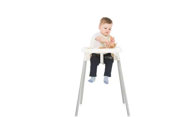 Little boy in bib eating bagels and sitting in highchair isolated on white background — Stock Photo