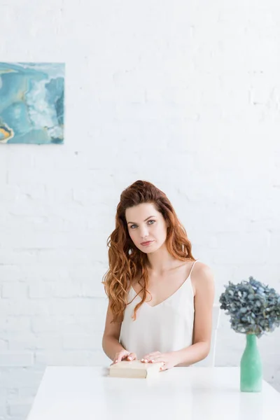 Beautiful young woman with book sitting at table and looking at camera — Stock Photo