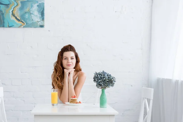 Bela jovem com suco de laranja e panquecas com morango para o café da manhã em casa olhando para a câmera — Fotografia de Stock
