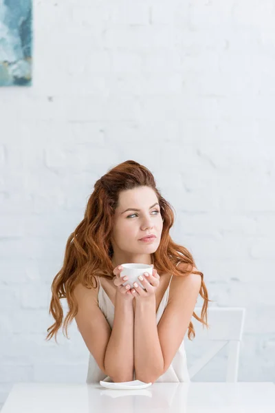 Thoughtful young woman with cup of coffee in front of white brick wall looking away — Stock Photo