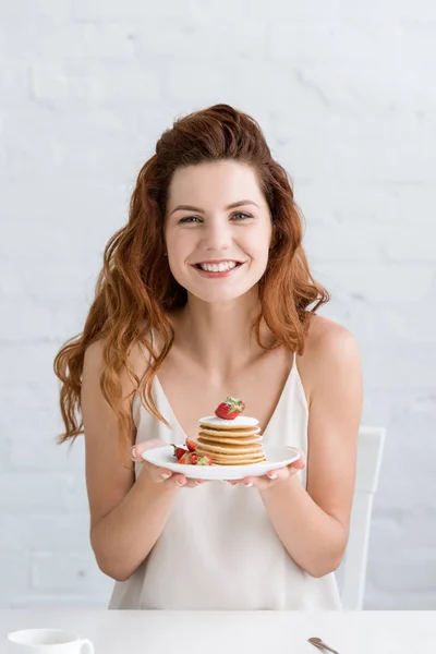 Schöne junge Frau hält leckere Pfannkuchen auf dem Teller und schaut in die Kamera — Stockfoto