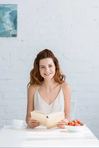 Sorrindo jovem mulher sentada à mesa com café e morango e livro de leitura em casa — Fotografia de Stock