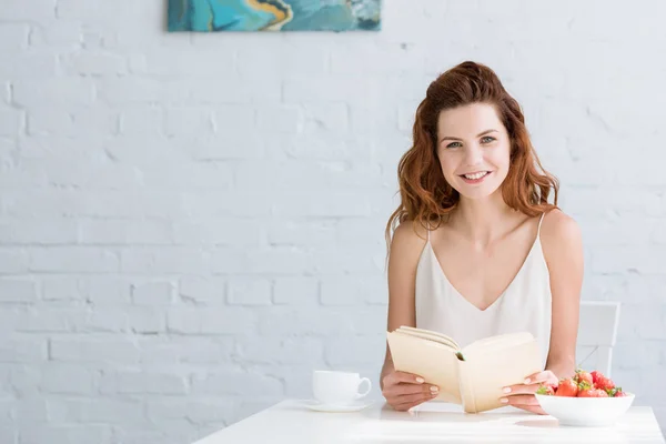 Happy young woman sitting at table with coffee and strawberry and reading book at home — Stock Photo