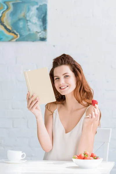 Feliz joven con libro y fresa sentado a la mesa - foto de stock