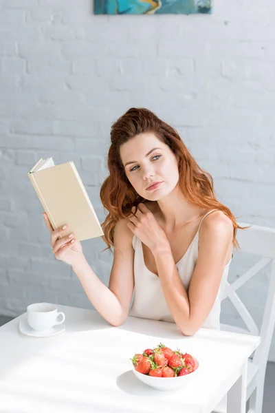 Mujer joven reflexiva con libro y taza de café sentado en la mesa - foto de stock