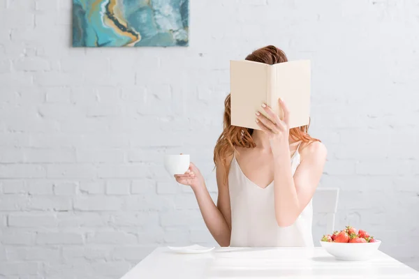 Mulher com xícara de café e tigela de morangos cobrindo rosto com livro em casa — Fotografia de Stock