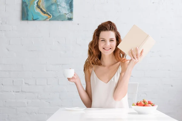 Heureuse jeune femme avec livre et tasse de café en regardant la caméra à la maison — Photo de stock