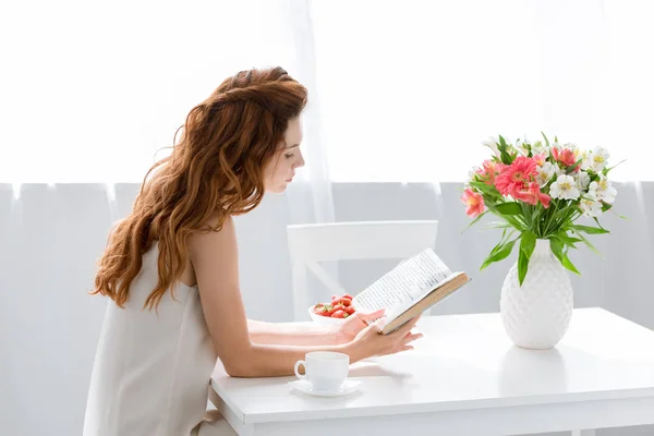 Mujer joven enfocada leyendo libro mientras está sentada en la mesa con taza de café y flores en jarrón - foto de stock