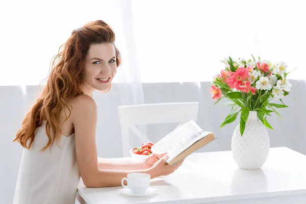 Felice giovane donna lettura libro mentre seduto a tavola con tazza di caffè e fiori in vaso — Foto stock