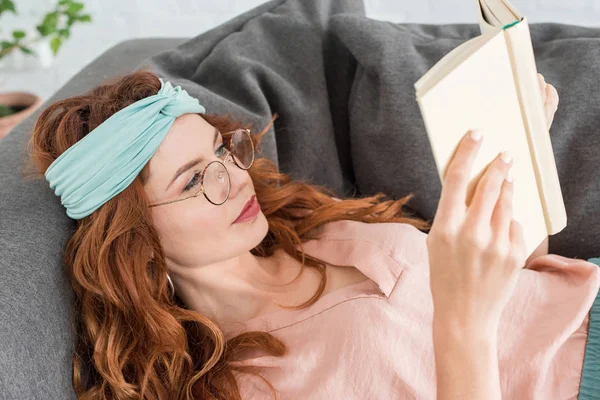 Schöne junge Frau liest Buch, während sie es sich zu Hause auf der Couch gemütlich macht — Stockfoto