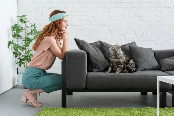 Smiling young woman looking at her tabby cat while he sitting on couch at home — Stock Photo