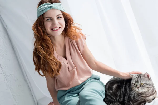 Smiling young woman petting adorable tabby cat while sitting on windowsill — Stock Photo