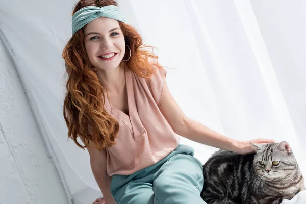 Sorrindo jovem mulher petting adorável gato tabby enquanto sentado no peitoril da janela — Fotografia de Stock