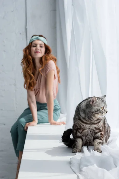 Happy young woman sitting on windowsill with tabby cat on foreground — Stock Photo