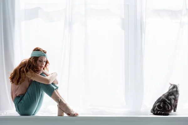 Mujer joven y solitaria sentada en el alféizar de la ventana con gato tabby - foto de stock