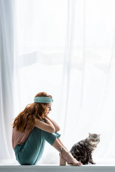 Réfléchie jeune femme assise sur le rebord de la fenêtre avec chat tabby — Photo de stock