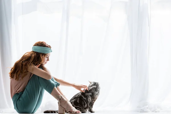 Hermosa joven mujer acariciando adorable tabby gato mientras sentado en windowsill - foto de stock