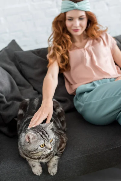 Belle jeune femme caressant chat tabby tout en étant assis sur le canapé à la maison — Photo de stock