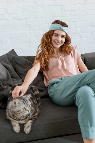 Lächelnde junge Frau streichelt gestromte Katze, während sie zu Hause auf der Couch sitzt — Stockfoto