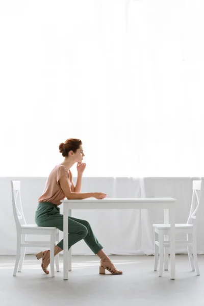 Vista lateral da bela jovem sentada à mesa sozinha em casa — Fotografia de Stock