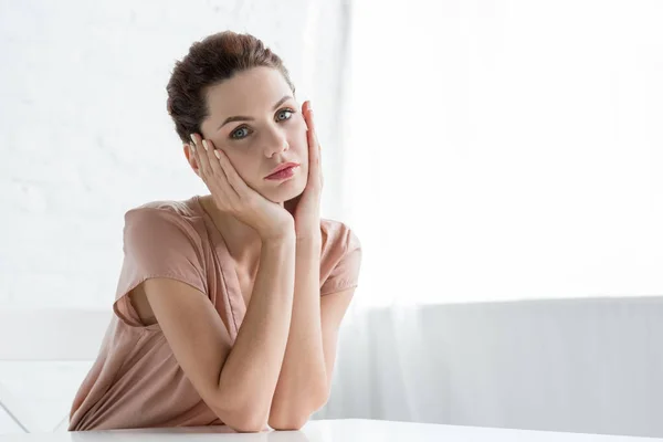 Nachdenkliche junge Frau sitzt am Tisch vor weißer Backsteinwand und blickt in die Kamera — Stockfoto