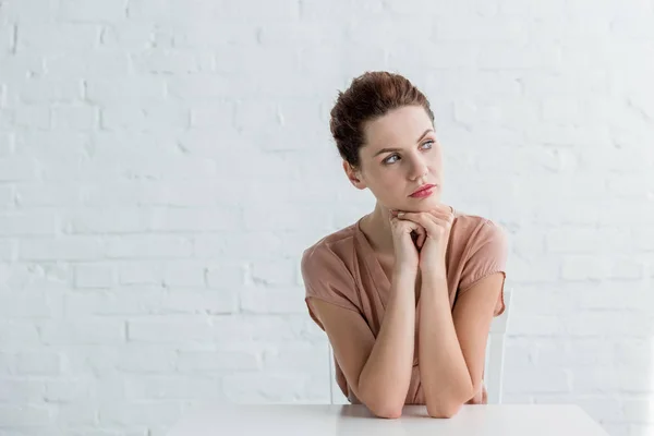 Mujer joven pensativa sentada a la mesa frente a la pared de ladrillo blanco — Stock Photo