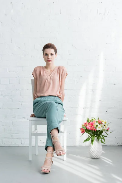 Attractive young woman sitting on chair with flowers on floor in front of white brick wall — Stock Photo